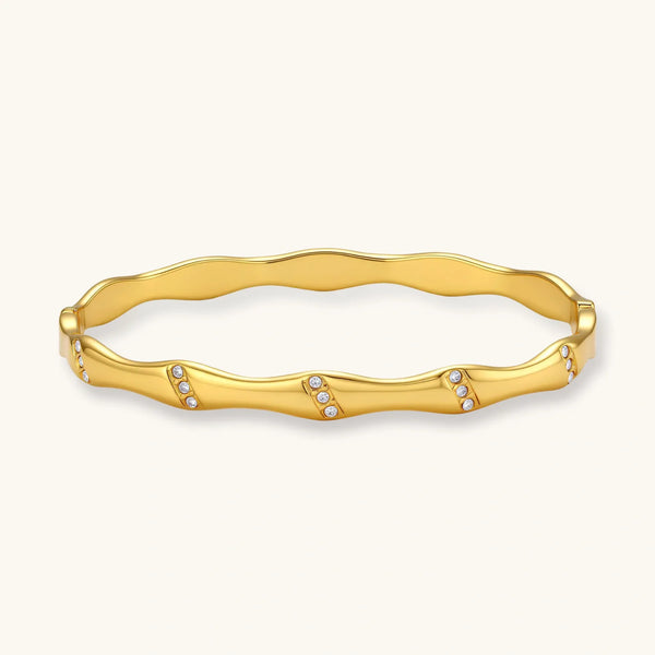 Gold-toned wavy bangle with bamboo-style segments and CZ crystal clusters on cream background. 18K plated stainless steel.